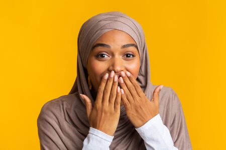 Happy Shock. Amazed black muslim girl in headscarf covering her mouth with hands over yellow background with free space, closeupの写真素材