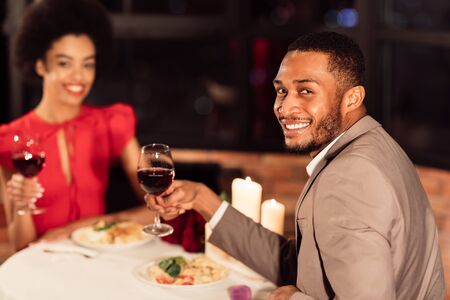 Romantic Dinner. Cheerful Black Couple Holding Red Wine Glasses Dating Smiling To Camera Celebrating...