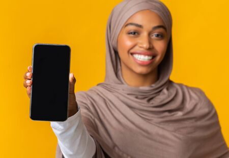 Modern smartphone with blank black screen in hand of smiling afro muslim girl in hijab, yellow background with empty spaceの写真素材