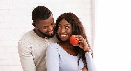 Happy afro woman with apple looking at her husband, man hugging woman from behind, white background, panorama with free spaceの写真素材