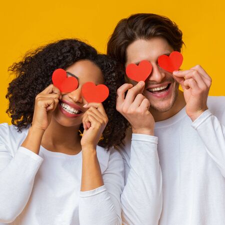 Closeup portrait of romantic interracial couple holding red paper hearts over eyes and sinserely...
