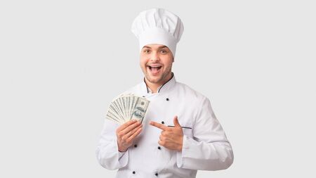 Restaurant Business. Excited Chef Man Holding Money Pointing Finger At Cash In His Hands Over White Background. Panoramaの写真素材