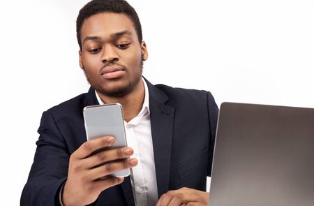 Chatting Online. Serious black businessman checking social media on smart phone, working on laptop,...
