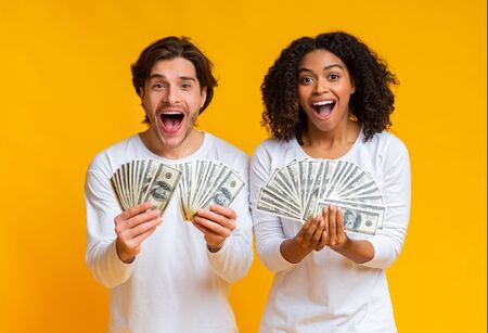Big Win. Overjoyed interracial couple holding lot of dollar cash and exclaiming with joy, celebrating success together, posing over yellow backgroundの写真素材