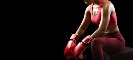 Female boxing concept. Cropped photo of female african boxer before fight, sitting on puff over black background, panorama with free spaceの写真素材
