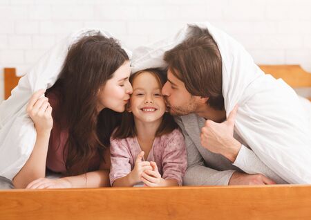 Family love. Happy parents kissing their little daughter in cheeks while hiding under blanket on bed, having fun together at homeの写真素材