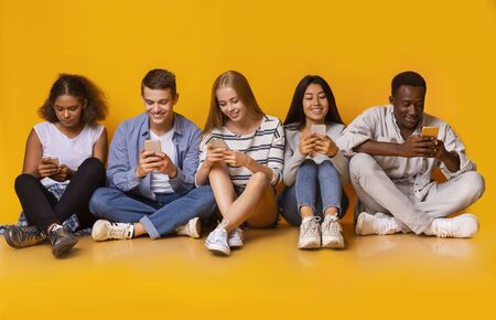Multiracial teenagers sitting on floor and texting with their smartphones, yellow background,...