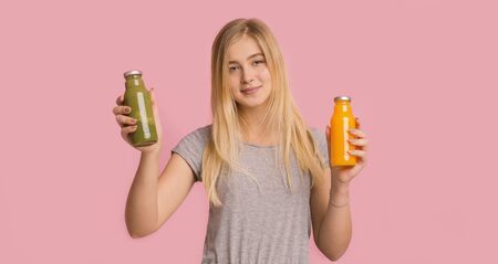 Sports nutrition. Smiling girl holding bottles with green and orange detox drink on pink background, panoramaの写真素材