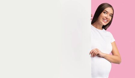 Happy Pregnant Woman Peeking Out Of Blank White Board For Text Advertising Something Standing Over Pink Studio Background. Mockupの写真素材