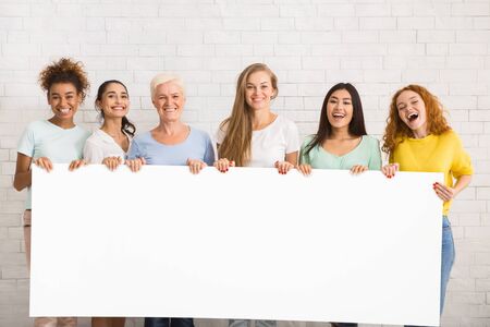 Multiracial Women And Girls Holding Empty White Poster For Text Smiling To Camera Standing Over White Wall. Mockupの写真素材