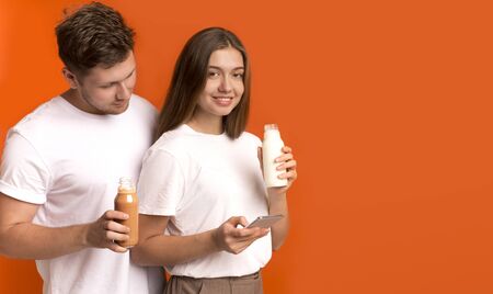 Healthy nutrition. Young couple drinking detox smoothies, girl using cellphone, orange background,...