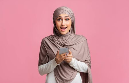Cool app. Excited muslim girl in hijab holding smartphone and looking to camera, enjoying new...