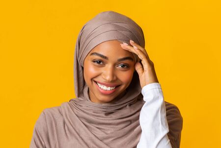 Closeup portrait of smiling afro muslim girl in headscarf shyly looking at camera and touching her...