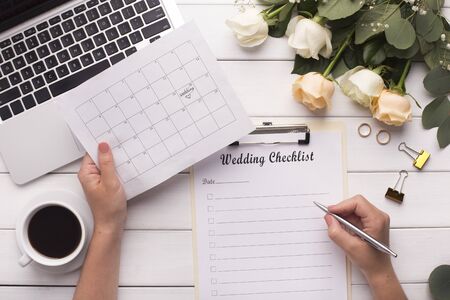 Bride Planning wedding writing checklist and choosing date in calendar, wooden background with cream colored roses and laptopの写真素材