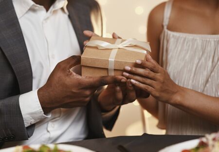 Valentine's surprise. Cropped of african man and woman holding gift box, having dinner at restaurantの写真素材