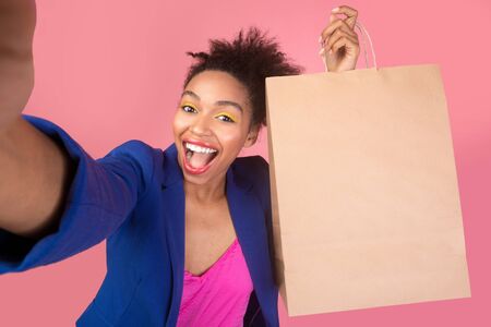 Winter Holiday Sale. Stylish afro woman taking selfie with shopping bag over pink studio wall, mock up, copyspaceの写真素材