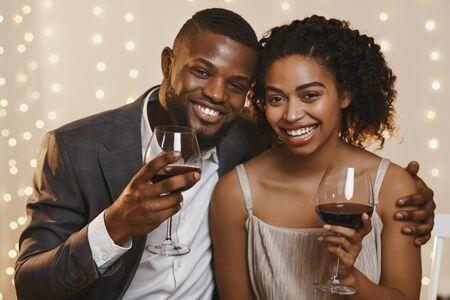 Portrait of cheerful african couple celebrating St. Valentines Day at restaurant, drinking red wine and embracing, close upの写真素材