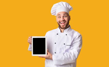 Cooking Food App. Joyful Male Chef Holding Digital Tablet Showing Blank Screen Standing Over Yellow Studio Background. Mockupの写真素材