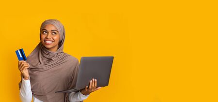 Online trading concept. Doubtful afro muslim girl in hijab holding credit card and laptop in hands, thinking about internet investment, panoramaの写真素材
