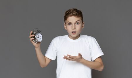 Education and time concept. Shocked teen boy holding alarm clock in hand, grey panorama backgroundの写真素材