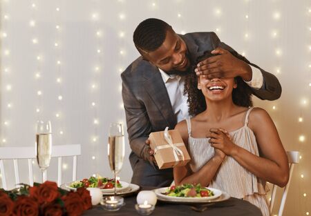 Black man in love making surprise to his excited woman on Valentine's Day, man closing woman eyes and holding gift box, restaurant interior, copy spaceの写真素材