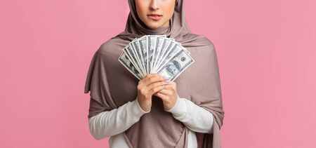 Money In Islam Concept. Doubtful arabic woman holding fan of dollar cash in hands, thinking about haram investment over pink background, panoramaの写真素材