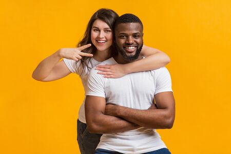 Cool Millennials. Portrait of cheerful afro guy and girl embracing and posing together on yellow background, young woman showing peace gestureの写真素材