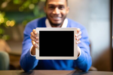 Cool App. Black young man showing tablet computer blank screen sitting at loft cafe, mock upの写真素材