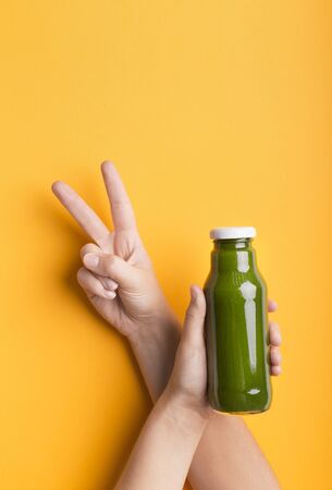 Cool and healthy. Girl showing v sign and advertising Glass bottle with green smoothie cocktail on yellow background, copy spaceの写真素材