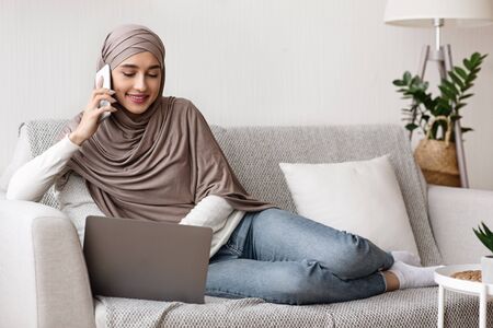 Muslim Girl In Hijab Talking On Phone And Using Laptop, Sitting On Couch at Homeの写真素材
