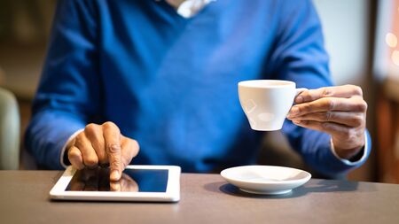 Young afro man sitting at the table with cup of coffee using digital tablet, man's hands typing...