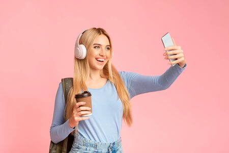Selfie Time. Portrait of blonde girl in headphones taking selfie using smartphone, holding cup of...