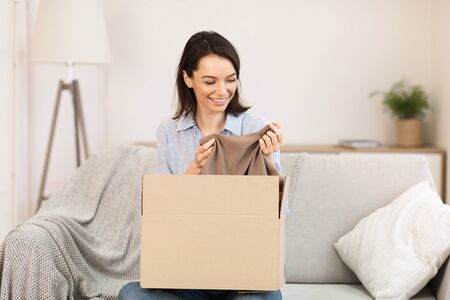 Buying Via Internet. Happy girl sitting on sofa unboxing cardboard delivery package after online...