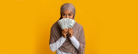 Happy Black Muslim Woman In Headscarf Covering Face With Dollar Cash Fan, Standing Over Yellow Background, Panorama With Copy Spaceの写真素材