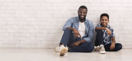 Like Father Like Son. Preteen black boy sitting on floor in similar pose with his dad near white brick wall, panorama with free spaceの写真素材