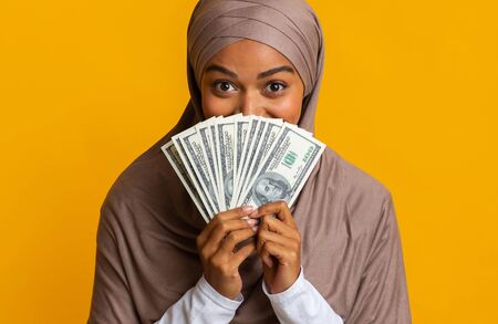 Great Bonus. Portrait Of Joyful Afro Islamic Woman In Hijab Hiding Face Behind Dollar Cash Fan In Her Handsの写真素材