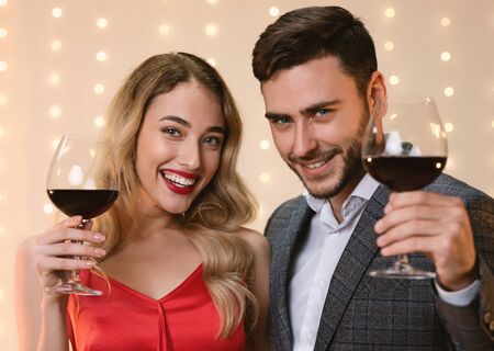 Cheers. Attractive Man And Woman Holding Glasses With Red Wine, Toasting And Smiling At Camera, Celebrating Holidaysの写真素材