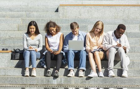 Multiethnic group of students preparing for exams, studying in park, using laptop, phone, tabletの写真素材
