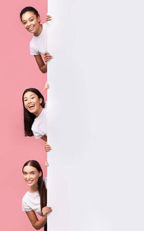 Great Offer. Three Multiracial Girls Peeking Out Of Blank White Board Posing Over Pink Studio Background. Mockup, Verticalの写真素材