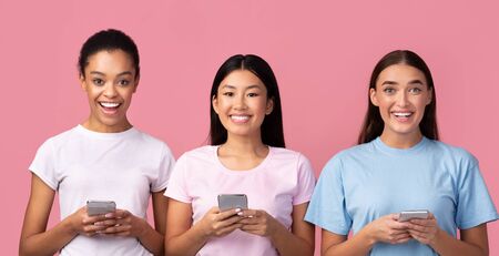 Three Happy Diverse Girls Using Smartphones Texting With Friends Standing Over Pink Studio...