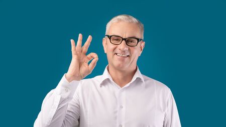 Everything is OK. Happy mature man in glasses showing approval gesture and smiling, blue studio...