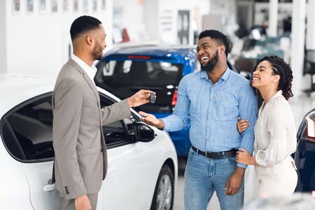 African American Couple Buying New Car Taking Key From Cheerful Salesman In Auto Dealership Showroom. Automobile Purchasing Serviceの写真素材