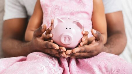 Afro father and little daughter holding pink piggy bank, close up, financial education at familyの写真素材