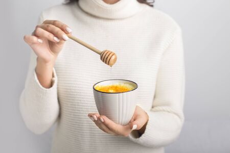 Female preparing turmeric latte drink with honey on gray background. Cup with yellow milk. Detox curcumin drinkの写真素材