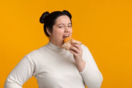 Gluttony Concept. Hungry Plus Size Girl With Overweight Eating Tasty Burger And Looking At Camera, Standing Over Yellow Background With Free Spaceの写真素材