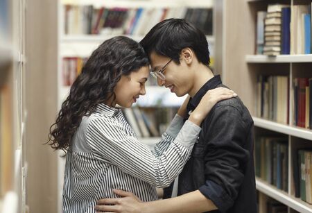 Date In Bookstore. Two shy multiethnic students hugging at library, flirting and sharing romantic...