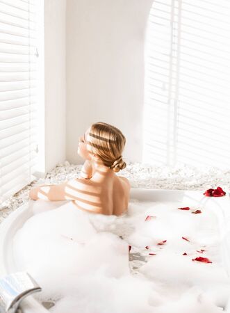 Back view of young blonde girl in sunlights enjoying foam bath at homeの写真素材