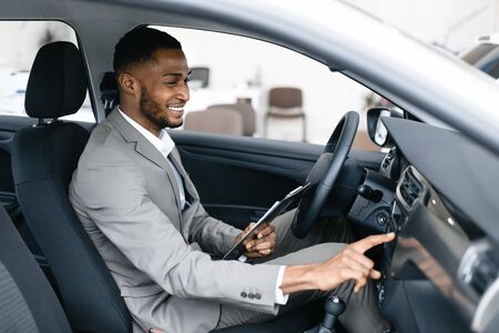Car Insurance. Afro Agent Man Checking Vehicle Taking Notes Sitting In Automobile In Dealership...