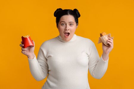 High Calorie Food. Shocked overweight girl holding hamburger and french fries pack in hands and looking at camera, yellow background, free spaceの写真素材