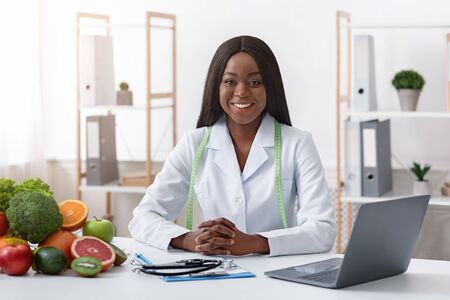 Smiling black lady doctor nutritionist sitting at workplace with vegetable and fruit, using laptop in officeの写真素材